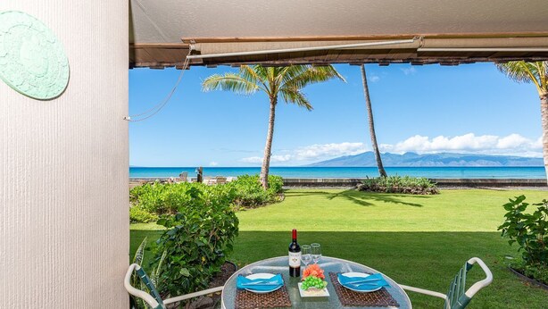 A table set for two with a bottle of wine, glasses, and a plate of food, overlooking a beach with palm trees and a clear sky.