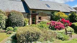 Uma casa de pedra com telhado inclinado, um pequeno alpendre e um jardim com arbustos e plantas floridas.