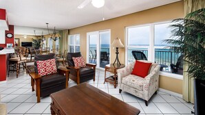 A spacious living area with a large wooden coffee table, multiple seating options including a sofa and chairs, a dining area with a TV, and large windows offering a view of the ocean. - IBR 7008: Memories by the Sea - Where Dreams Meet the Ocean! (Fort Walton Beach)