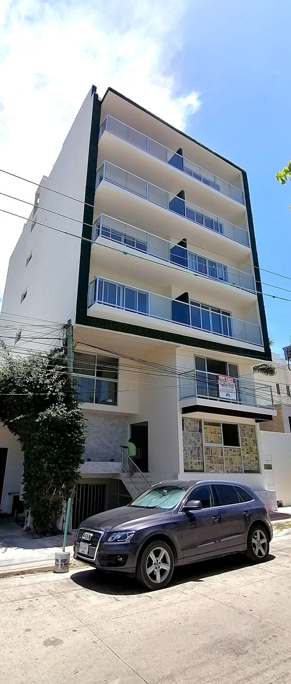 A multi-story building with balconies, a car parked in front, and a 'For Sale' sign.