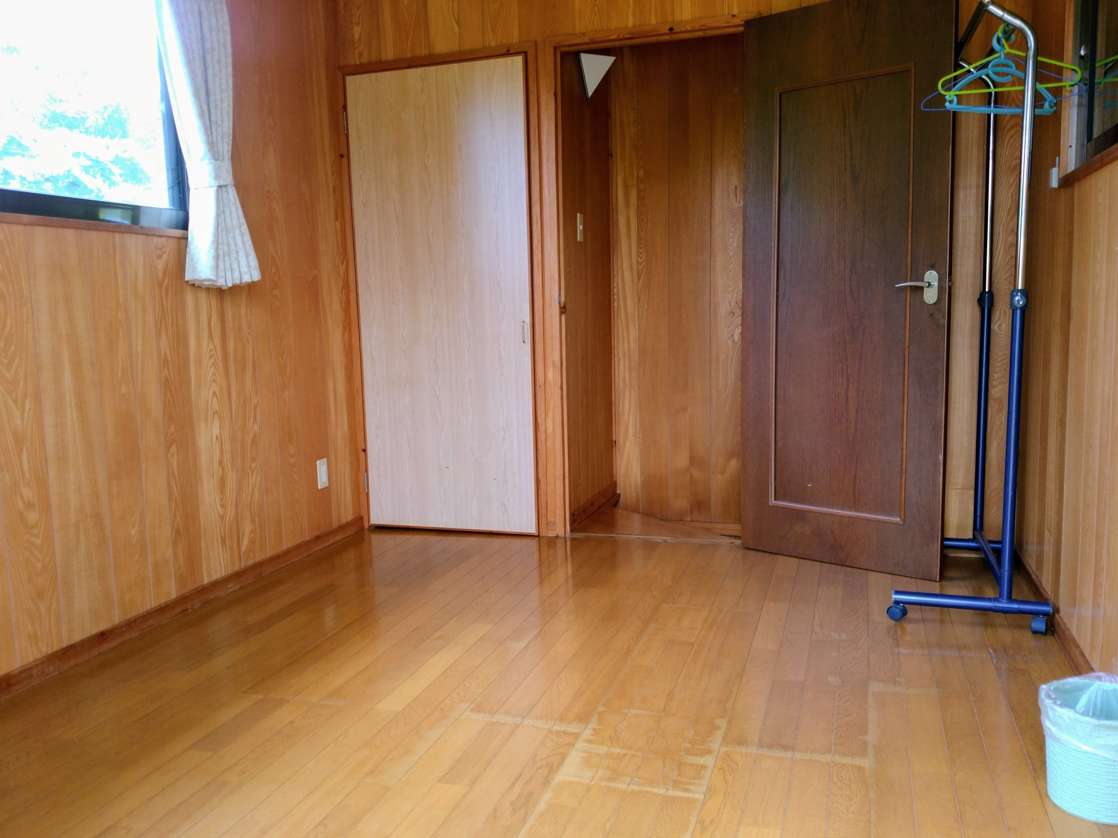 A wooden-paneled room with a wooden floor, a door, a coat rack, and a trash bin.