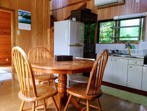 A wooden interior with a round wooden table and four chairs, a white refrigerator, a kitchen area with a window, and a calendar on the wall. - Cottage CANAC (Motobu)