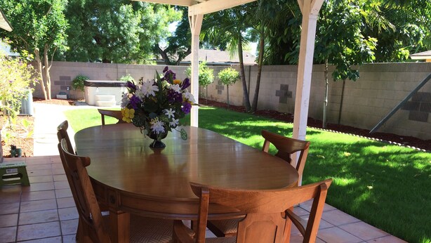 Een terras buiten met een houten tafel en stoelen, omgeven door een tuin en een bakstenen muur.