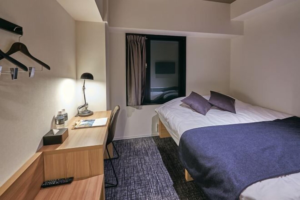 Une chambre d’hôtel compacte comprenant un lit simple, un bureau avec une lampe, une chaise, une petite table avec une télécommande et une fenêtre avec des rideaux.