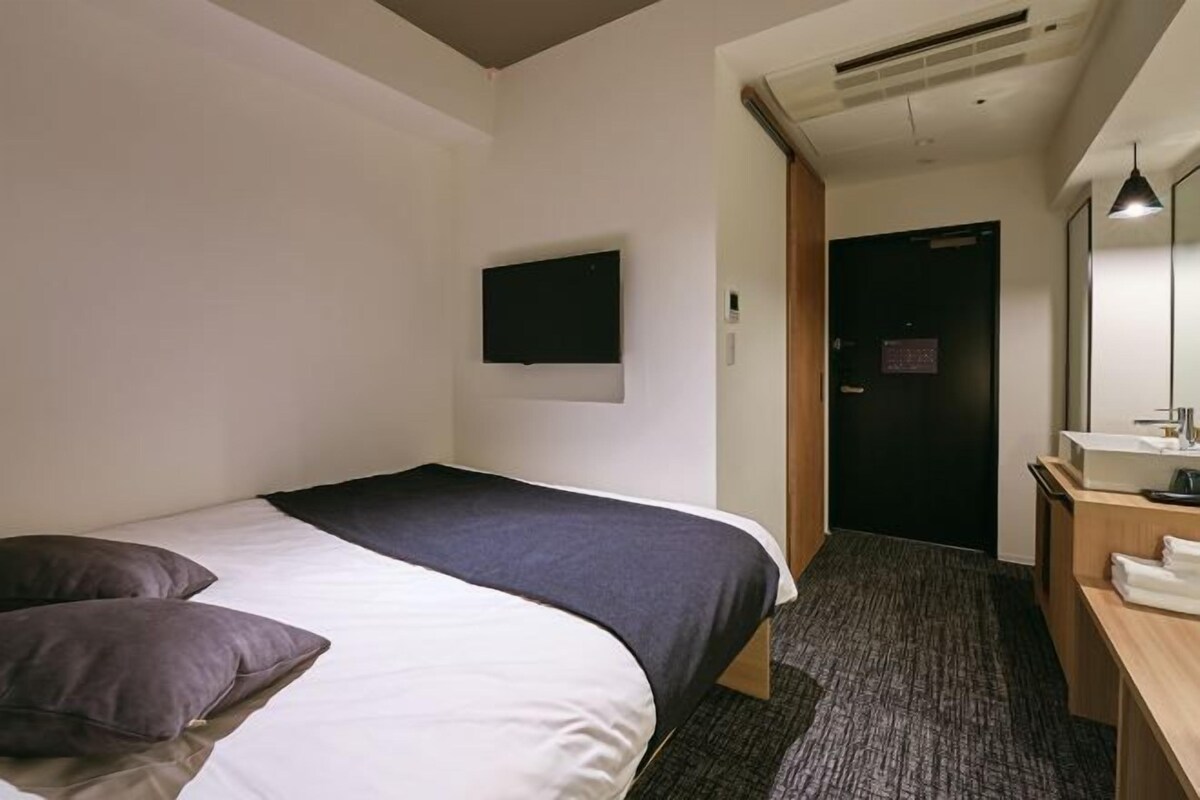 A hotel room with a bed, a flat-screen TV mounted on the wall, a desk with a mirror and a sink, and a door leading to another room.