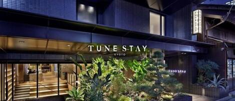 Una entrada de hotel moderna con un letrero que dice "TUNE STAY" y un interior bien iluminado visible a través de las puertas de vidrio.