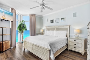 A bedroom with a bed, bedside tables, a ceiling fan, a TV, a plant, and a view of the ocean.