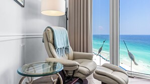 Ein Zimmer mit großem Fenster und Blick auf den Strand, ein beiges Sessel, ein Glastisch und eine Lampe.