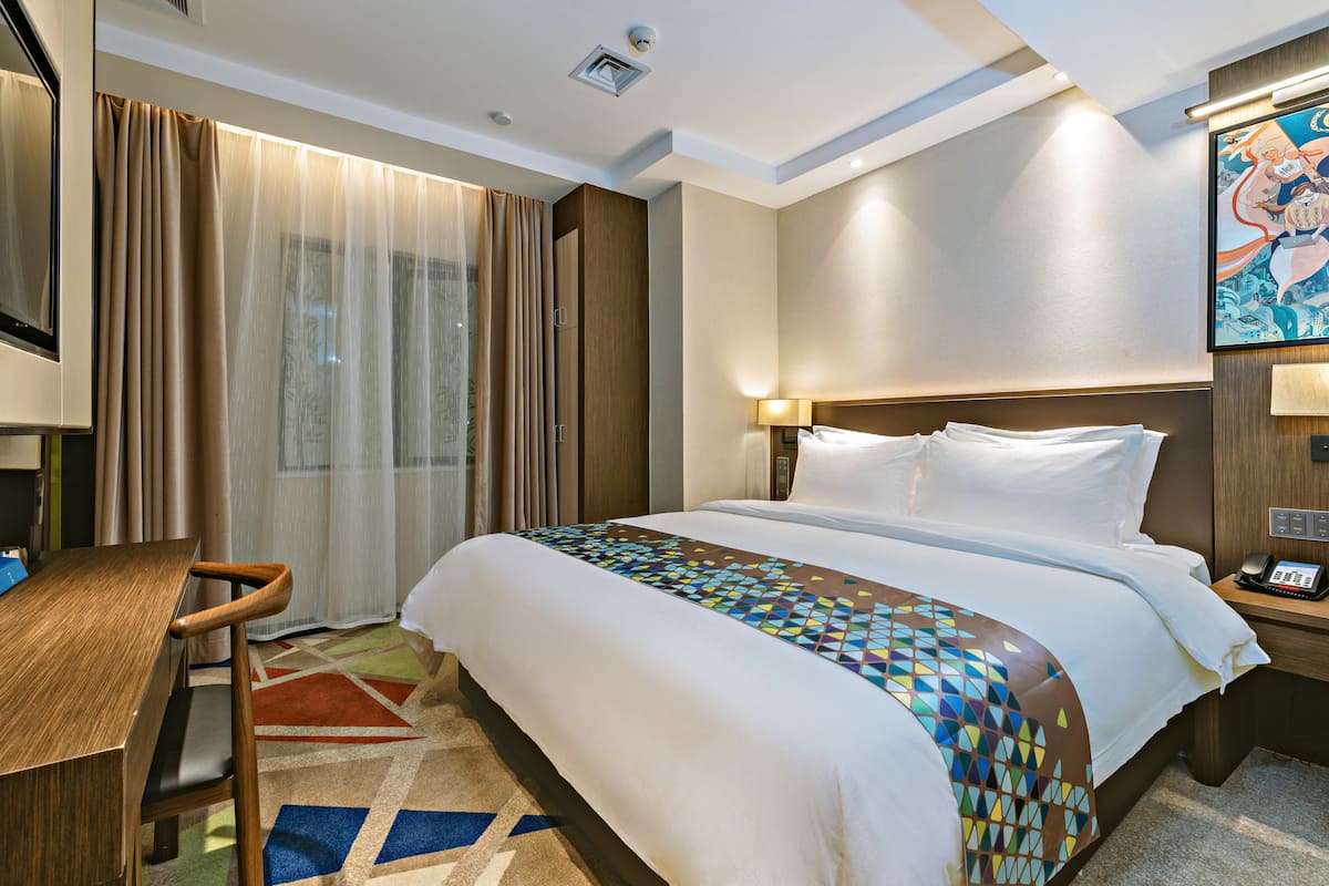 A hotel room with a large bed, a desk, and a chair. There is a window with curtains, a painting on the wall, and a lamp on the bedside table.