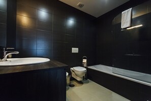 A modern bathroom with a dark tiled wall, a white sink, a toilet, and a bathtub.