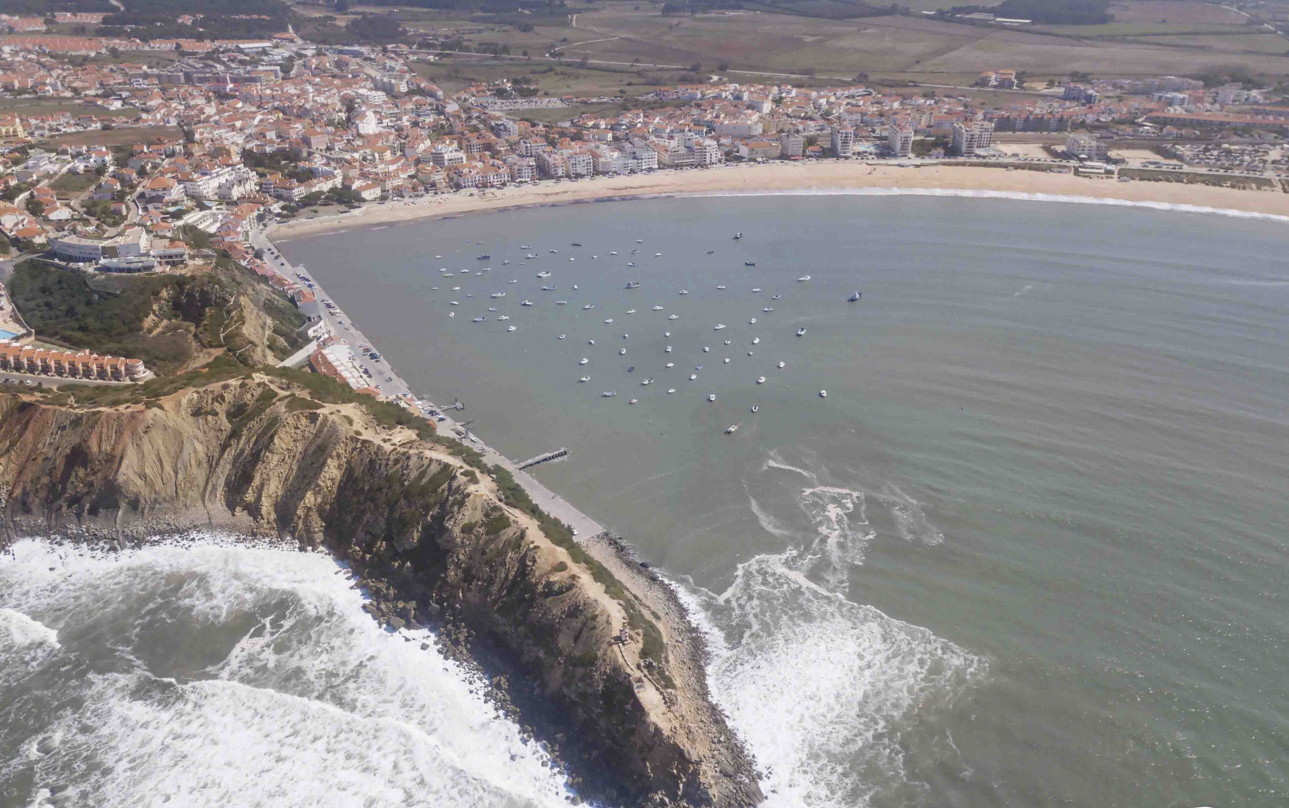 An aerial view of a coastal town with a beach, boats, and rocky cliffs.