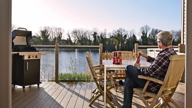 Ein Mann sitzt auf einem Liegestuhl an einem Tisch mit Grill und Flaschen und blickt auf einen See.
