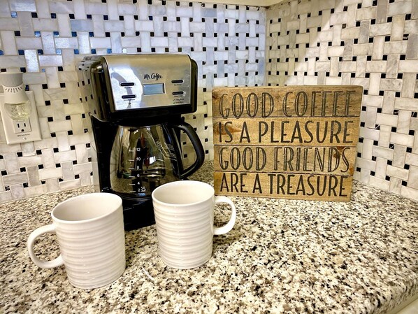 A coffee maker, two mugs, and a wooden sign with a coffee-related message.