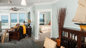 A spacious living room with a large window overlooking the ocean, a ceiling fan, and a bookshelf.