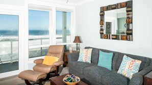 A living room with a view of the ocean, a leather chair, a gray sofa with patterned cushions, and a decorative mirror.