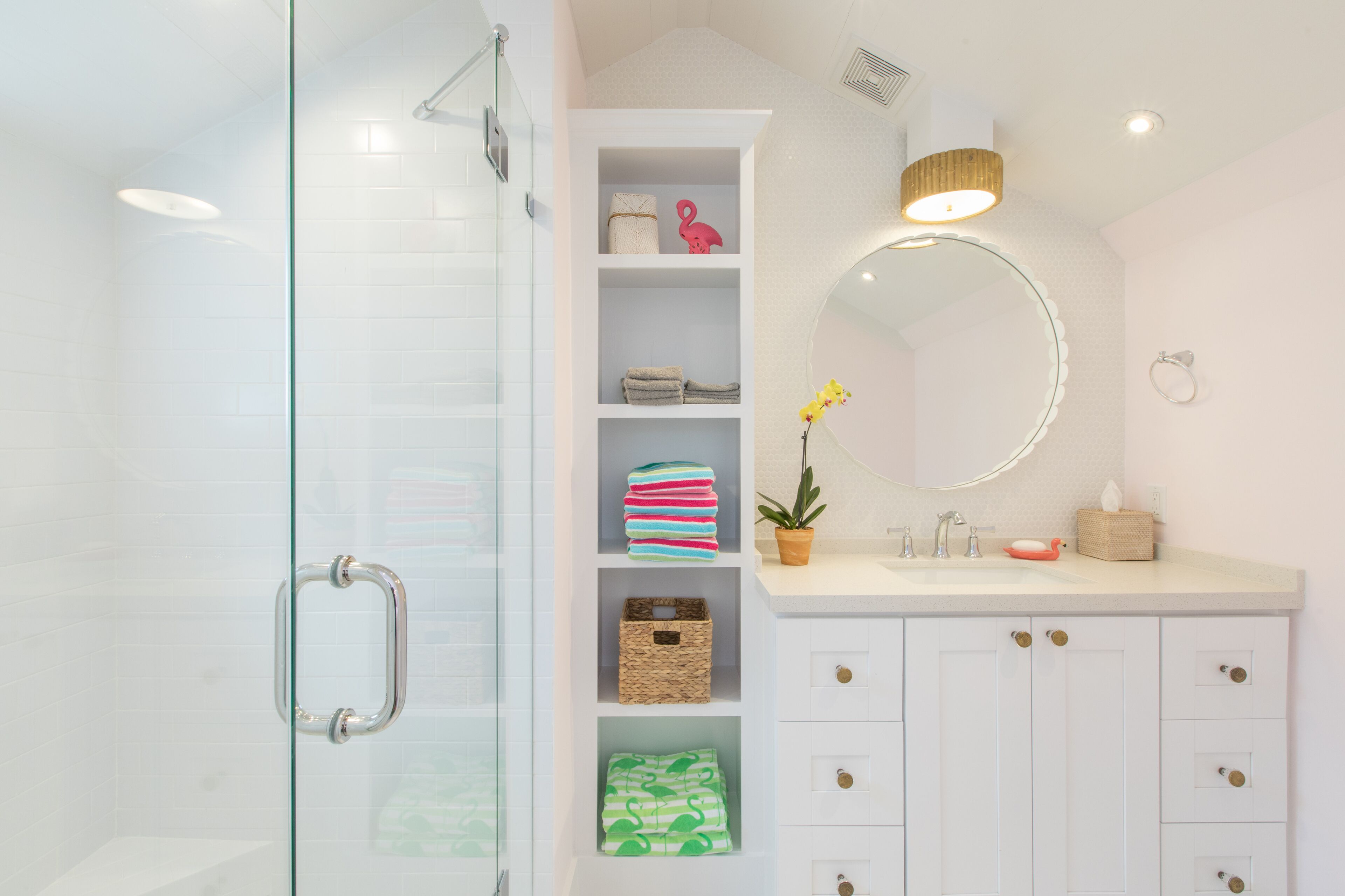 A bathroom with a glass shower enclosure, white cabinets, a vanity with a mirror, and shelves with towels and toiletries.