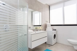 A modern bathroom with a glass shower enclosure, a white sink with a mirror, a washing machine, and a toilet. - Stylish Spacious 3BR Apartment 5min From the Beach (Tel Aviv-Yafo)