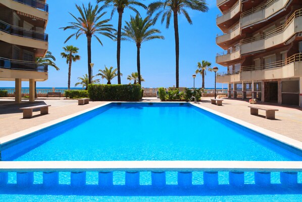 A swimming pool with a clear blue water surface, surrounded by palm trees and a building with balconies. - Beachfront Apartment with Pool · UHC SOL DE ESPAÑA 208 (Cambrils)