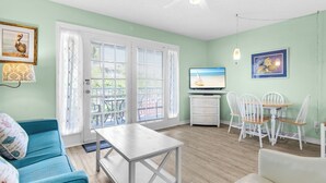 A living room with a teal sofa, a white coffee table, a TV on a stand, a dining table with chairs, and a ceiling fan. - At the Beach - Gulf Views - Pool (Miramar Beach)