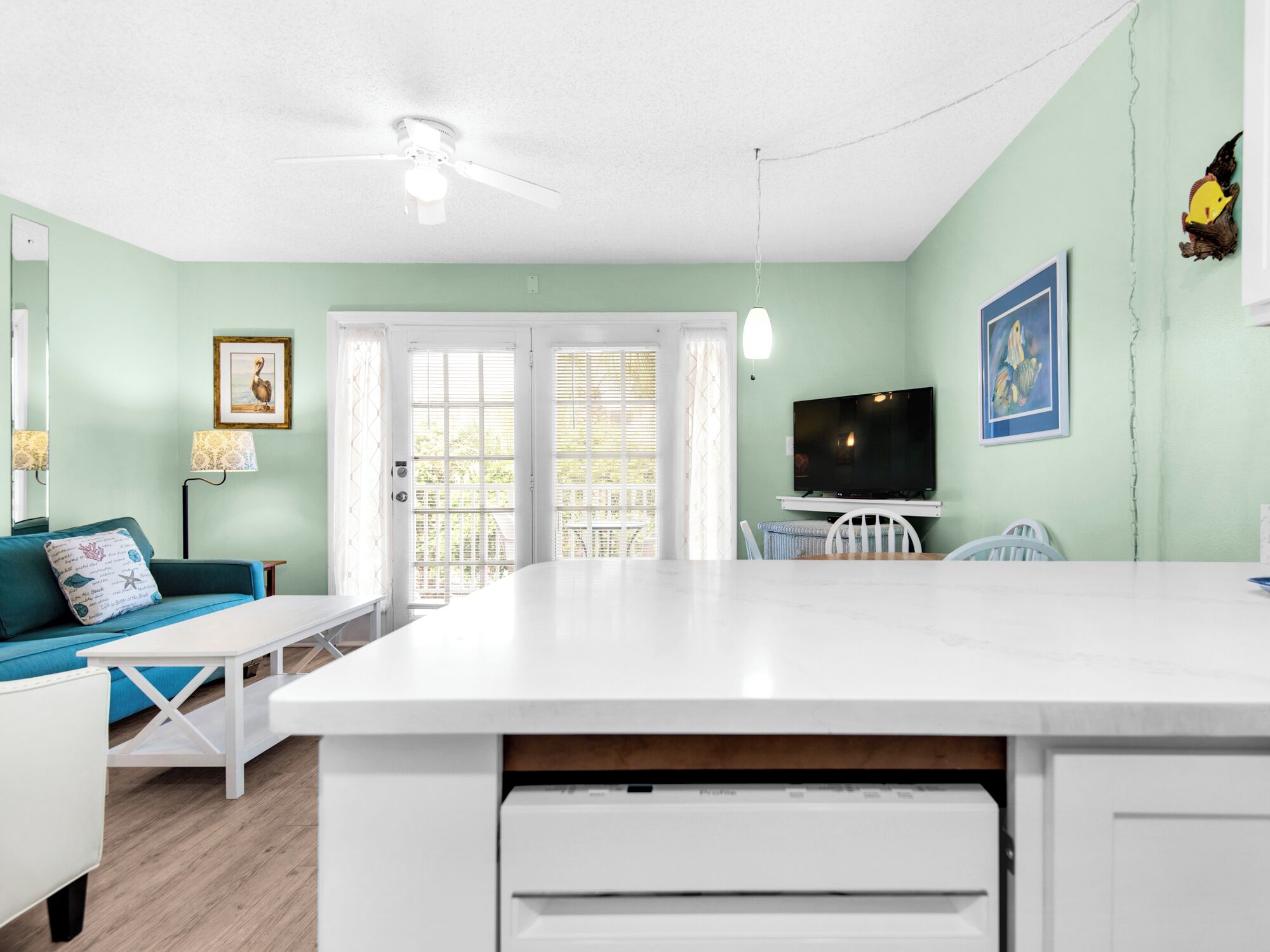 Una habitación luminosa y bien iluminada, con un ventilador de techo blanco, un ventanal con vistas, un sofá color turquesa con un cojín decorativo, una mesa de comedor blanca y un televisor sobre un mueble bajo.