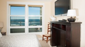 A hotel room with a large window offering a view of the ocean, a bed with patterned bedding, a wooden chair, a dark wooden cabinet, and a flat-screen TV mounted on the wall. - Promo! Oceanfront Condo w/ Private Hot Tub, Indoor Pool, WiFi and More! (Lincoln City)