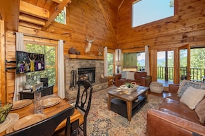 A wooden cabin interior with a stone fireplace, a television, a dining table, and a cozy seating area. - AMAZING VIEWS! Firepit, Private, Woodburning Fireplace, Hot Tub, Pool Table! (Sevierville)