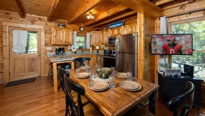 A wooden cabin with a kitchen featuring a dining table set for four, a refrigerator, microwave, and a television displaying a football game. - AMAZING VIEWS! Firepit, Private, Woodburning Fireplace, Hot Tub, Pool Table! (Sevierville)