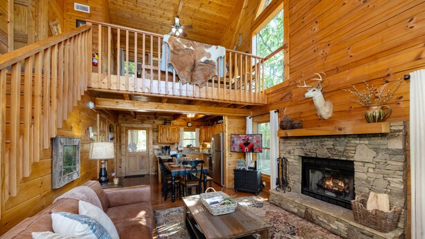 A spacious log cabin interior with a high ceiling, a stone fireplace, a dining area, and a cozy living room with a large leather sofa. - AMAZING VIEWS! Firepit, Private, Woodburning Fireplace, Hot Tub, Pool Table! (Sevierville)