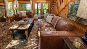 A cozy log cabin interior with a leather sofa, a coffee table, and a large window overlooking a forest. - AMAZING VIEWS! Firepit, Private, Woodburning Fireplace, Hot Tub, Pool Table! (Sevierville)
