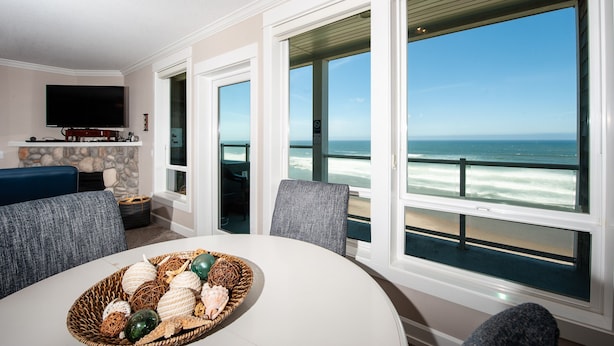 A room with a large window overlooking the ocean, a dining table with a basket of decorative items, and a flat-screen TV mounted on the wall. - Promo! Second Floor Corner Condo, Oceanfront, Private Hot Tub, Pool, Wifi (Lincoln City)