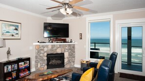 A living room with a stone fireplace, a television, a bookshelf, and a view of the ocean through a large window. - Promo! Second Floor Corner Condo, Oceanfront, Private Hot Tub, Pool, Wifi (Lincoln City)