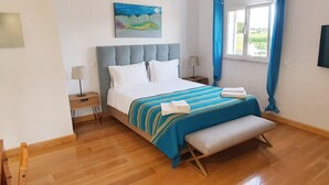 Een slaapkamer met een bed, nachtkastje, lamp, bankje en een schilderij aan de muur.