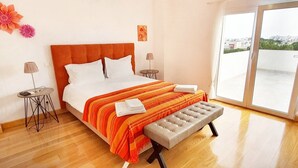 Een moderne slaapkamer met een groot bed, een bankje, twee nachtkastjes en uitzicht op de stad door de balkondeuren.