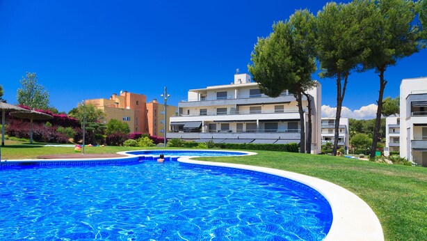 A swimming pool with a curved edge, surrounded by grass and trees, with buildings in the background. - Penthouse with pool view - UHC SALOU VILLA FAMILY COMPLEX 029 (Salou)