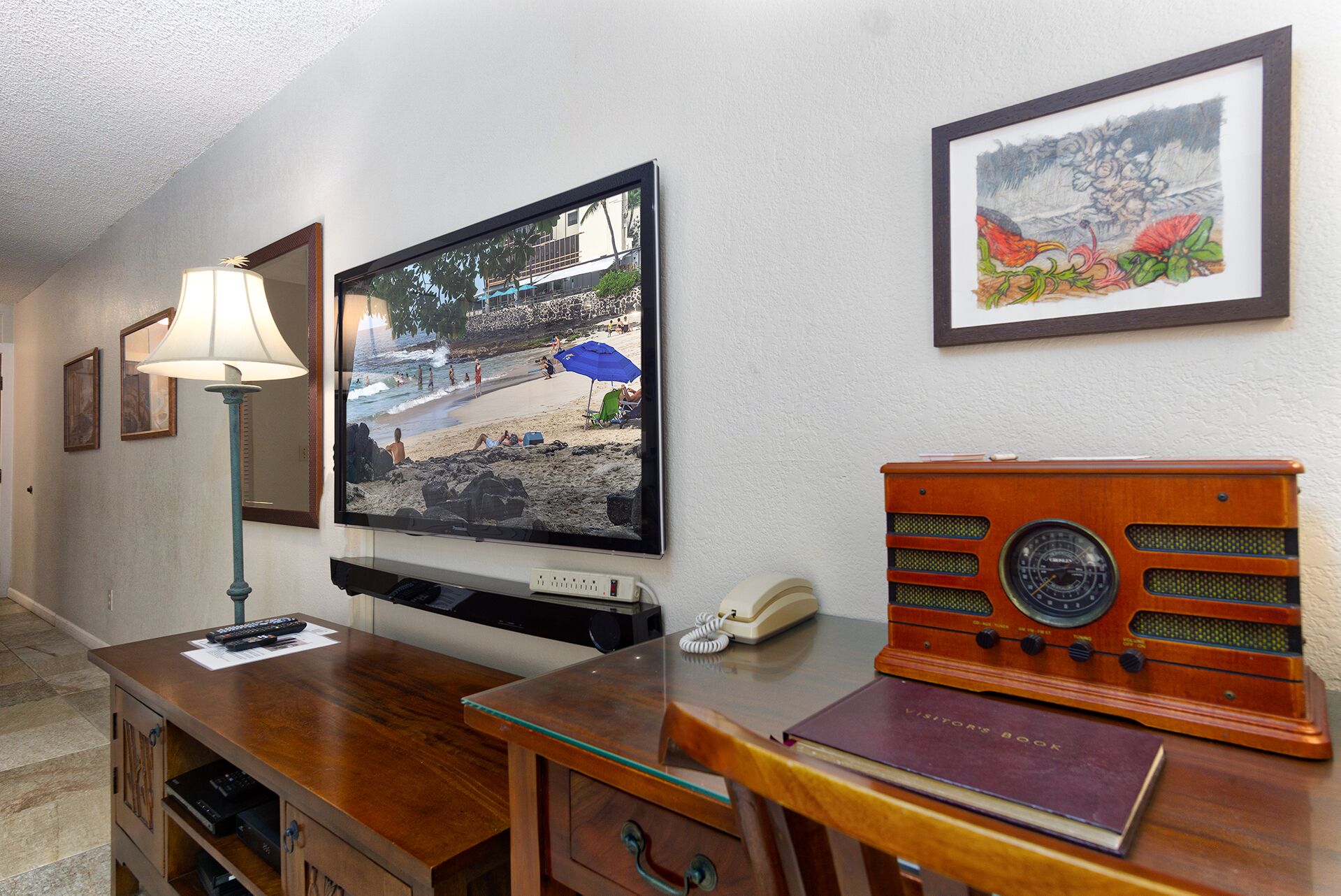 A hotel room with a television mounted on the wall, a wooden desk with a telephone and a book, and a framed picture above the desk.