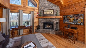Interior acogedor de una cabaña con chimenea de piedra, paredes de madera, un ventanal grande y un televisor.
