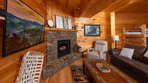 Un interior de cabaña acogedor con chimenea de piedra, paredes de madera, un sofá cómodo y un televisor.