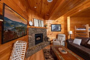 Ein gemütliches Blockhaushaus-Interieur mit Kamin aus Naturstein, Holzwänden, einer bequemen Couch und einem Fernseher.