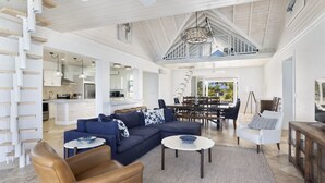 A spacious living area with a large sectional sofa, a brown leather chair, and a dining area with a high ceiling and a staircase. - Compound w/Beach, Pool,  Pickleball, Gym, Kayaks, Golf Carts, Views! (Governors Harbour)