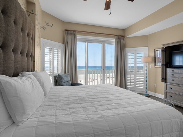 A large bed with white bedding, a ceiling fan, and a view of the beach through the balcony doors.