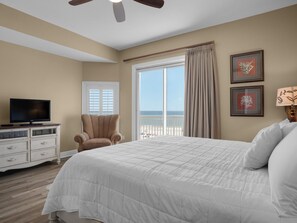 A bedroom with a large bed, a chair, a TV, and a window with a view of the beach.