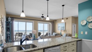 A kitchen with a granite countertop, stainless steel sink, and white cabinetry. There is a ceiling fan, pendant lights, and a view of the ocean through large windows. - Hear the Waves, Big Balcony, Look at the Beach, Unique, Spacious (Miramar Beach)
