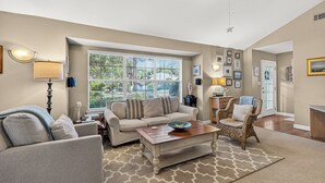Un salon comprenant un canapé, des fauteuils, une table basse et une chaise en osier. On y trouve une grande fenêtre avec des stores, une lampe et des cadres photos accrochés au mur.
