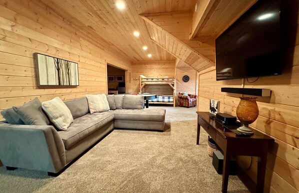 A cozy log cabin interior with a sectional sofa, a TV mounted on the wall, and a wooden ceiling.