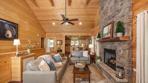 A cozy living room with a stone fireplace, a ceiling fan, and a view into the kitchen.
