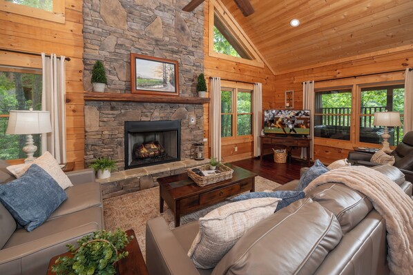 A log cabin interior with a stone fireplace, leather sofas, wooden floors, and large windows.