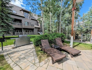 Une terrasse dotée de deux fauteuils de jardin en osier, d’un barbecue et d’un chemin de pierre menant à un bâtiment avec un balcon.