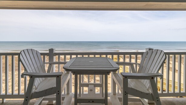 A deck with two Adirondack chairs and a small table, overlooking a beach with a wooden fence. - FFR310 - Ocean Sky by First Flight Rentals (Kill Devil Hills, NC 27948)