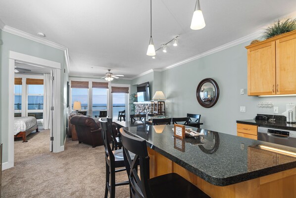 A spacious kitchen with a large island, a dining area with chairs, a ceiling fan, and a view of the ocean through large windows.