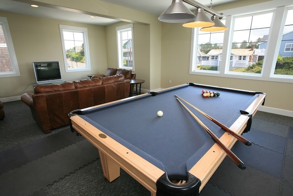 A pool table with a white cue ball, pool cues, and billiard balls. There is a brown leather couch and a television in the background.